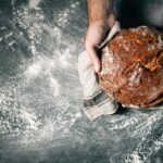 Hand haelt frisches Brot mit Nuesse auf dunkler bemehlter Arbeitsflaeche – Nussbrot selber backen mit Charakter.