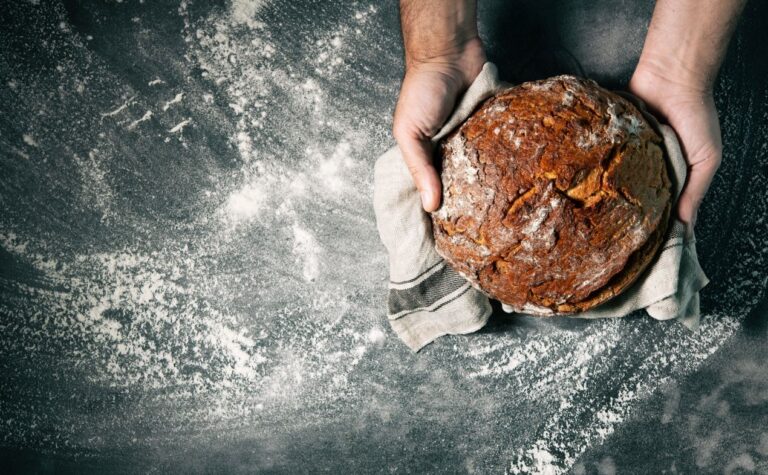 Hand haelt frisches Brot mit Nuesse auf dunkler bemehlter Arbeitsflaeche – Nussbrot selber backen mit Charakter.