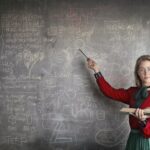 Frau schreibt etwas an die Tafel und guckt erwartungsvoll in die Kamera